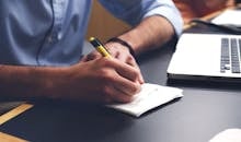 Close-up of a man writing notes with a pen next to a laptop on a desk.